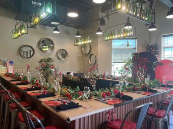 Indoor restaurant bar set for a birthday party with U-shaped counter, red rose garlands, wine glasses, black napkins, metal stools and green bottle chandeliers