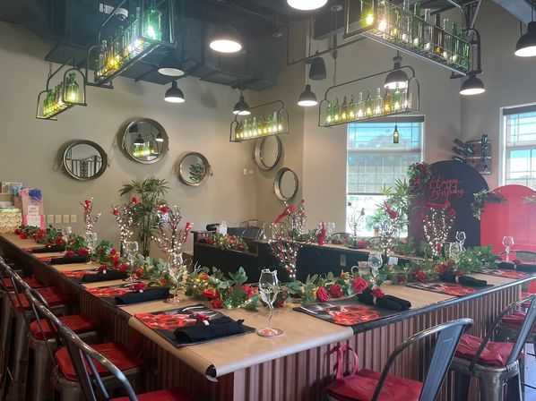 Indoor restaurant bar set for a birthday party with U-shaped counter, red rose garlands, wine glasses, black napkins, metal stools and green bottle chandeliers