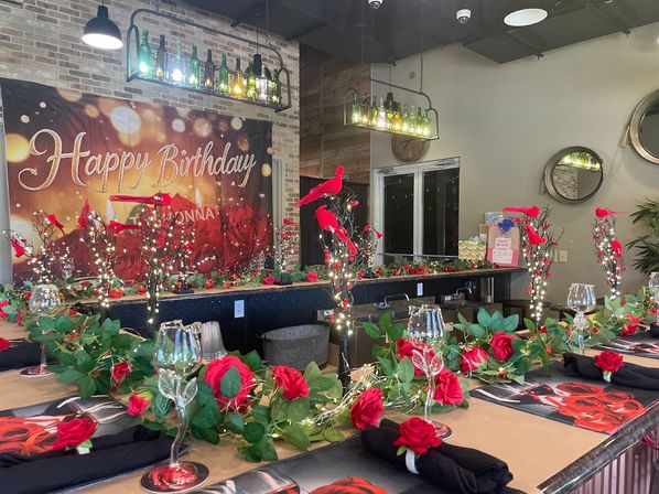 Indoor birthday setup in a bar-style venue with a large Happy Birthday banner, long table decorated with red rose garlands, faux red bird accents, twinkling fairy lights, wine glasses and bottle pendant lights.