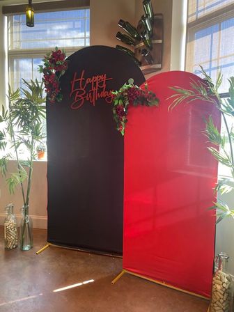Indoor birthday photo backdrop featuring arched black and red panels with a red "Happy Birthday" script, rose garlands, bamboo plants, wine bottle wall display and cork-filled glass vases in a sunlit event space.