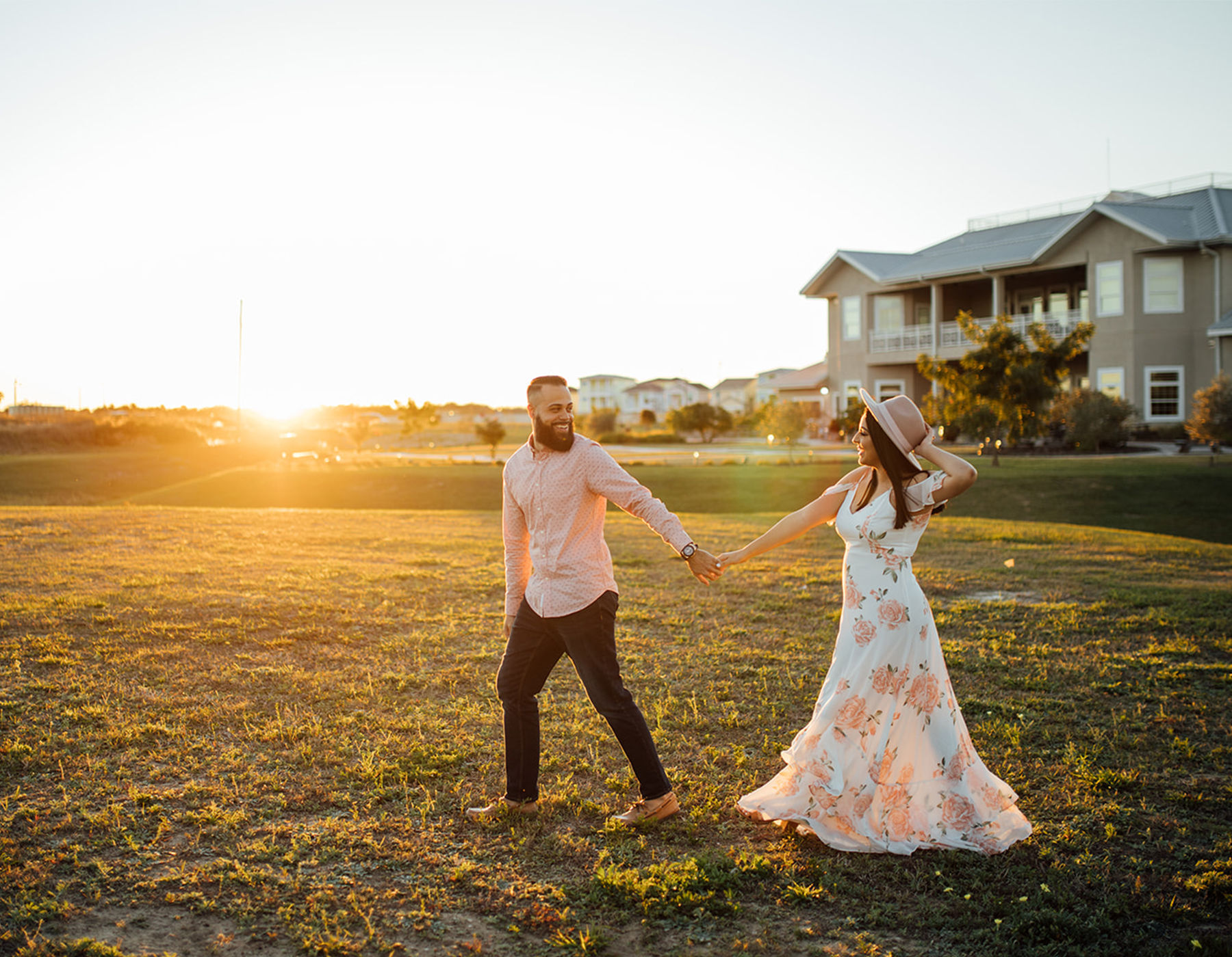 Couple enjoying a sunset stroll across a grassy field near suburban houses — woman in a floral maxi dress and hat holding hands with a bearded man in a casual shirt.