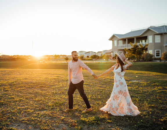 Couple enjoying a sunset stroll across a grassy field near suburban houses — woman in a floral maxi dress and hat holding hands with a bearded man in a casual shirt.