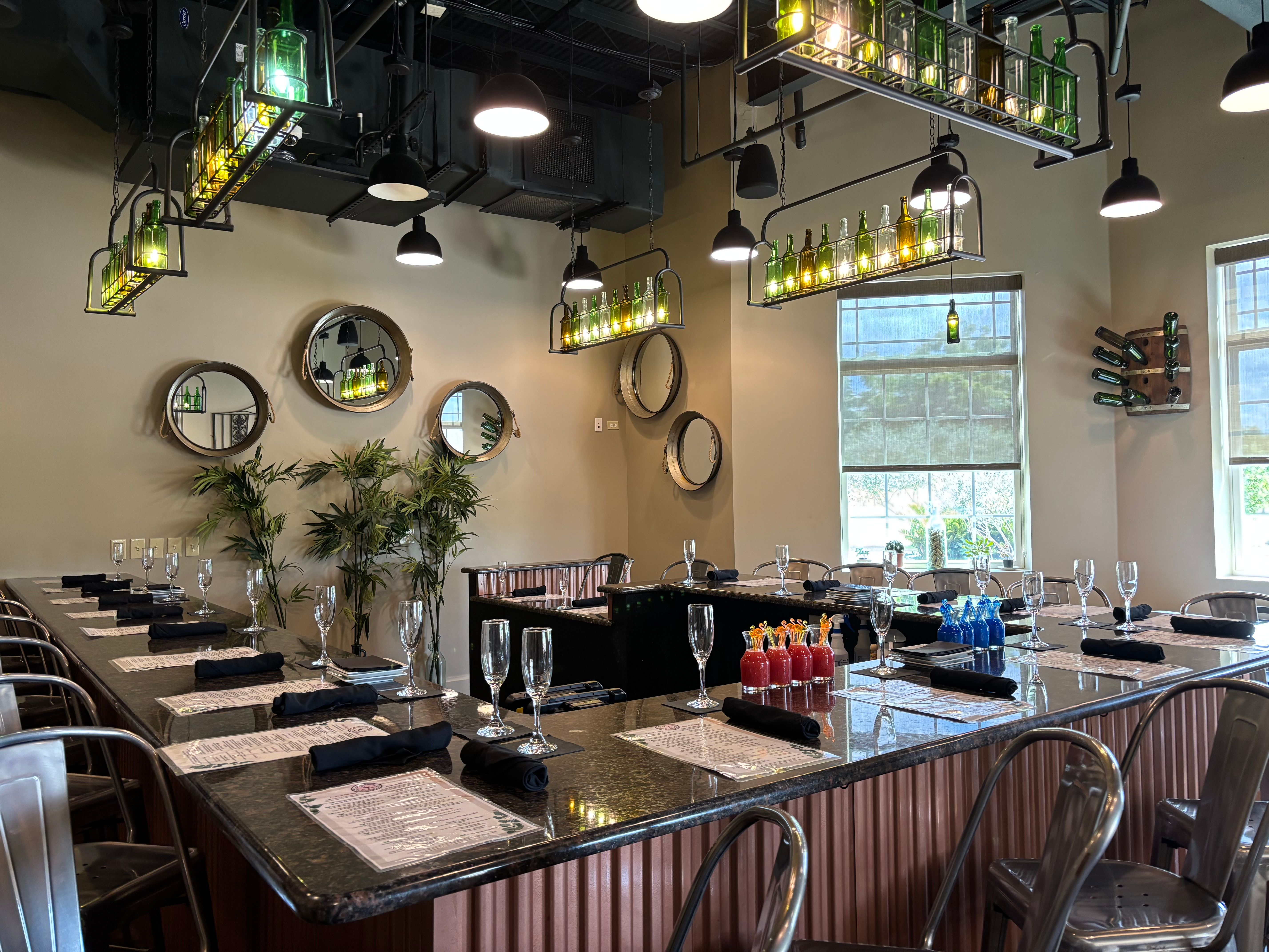 Modern industrial restaurant dining counter with U-shaped granite bar, metal stools, place settings with menus, black napkins and wine glasses, hanging green-bottle light fixtures, round wall mirrors and potted plants by sunlit windows