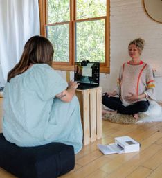 Pregnant woman practicing prenatal meditation in a sunlit home studio, seated cross-legged on a rug by a large window while another person runs a laptop on a wooden crate for a virtual session.