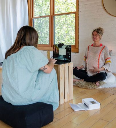 Pregnant woman practicing prenatal meditation in a sunlit home studio, seated cross-legged on a rug by a large window while another person runs a laptop on a wooden crate for a virtual session.