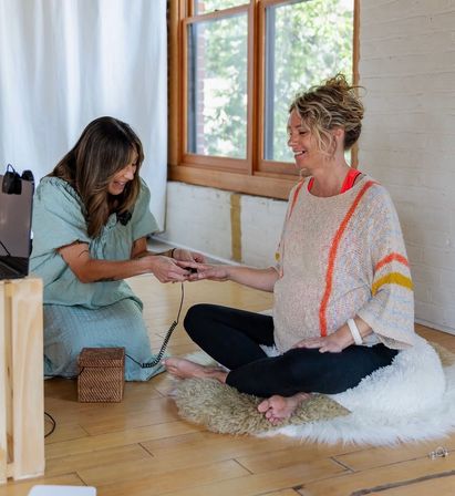 Cozy prenatal session in a sunlit wooden-floor studio: a smiling pregnant woman sits cross-legged on a rug while a practitioner hands her a small corded device, both relaxed and laughing.