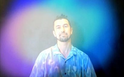 Artistic studio portrait of a man in a light-blue button-up shirt with a soft smile, bathed in vivid purple-to-teal gradient light and haze