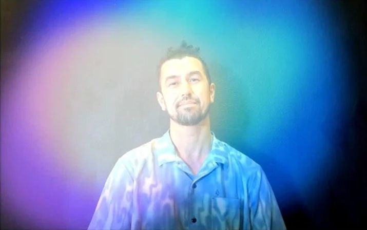 Artistic studio portrait of a man in a light-blue button-up shirt with a soft smile, bathed in vivid purple-to-teal gradient light and haze
