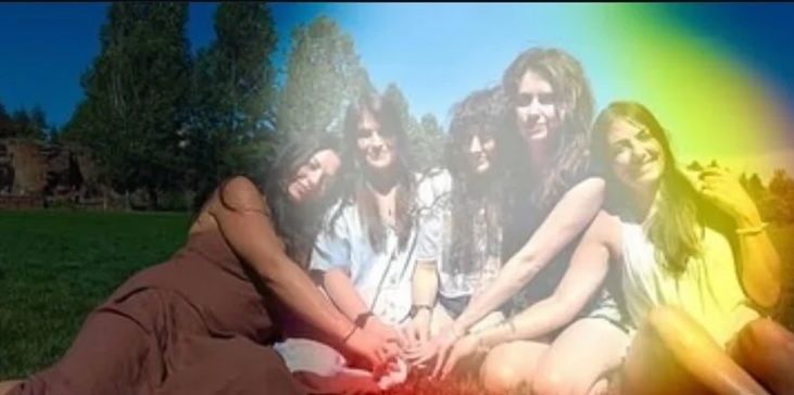 Five young women sitting on a green lawn in a sunny park, smiling and holding hands in a circle with tall trees behind them and a colorful rainbow light-leak across the photo.