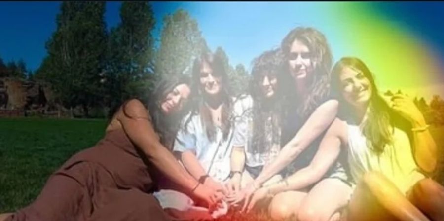 Five young women sitting on a green lawn in a sunny park, smiling and holding hands in a circle with tall trees behind them and a colorful rainbow light-leak across the photo.