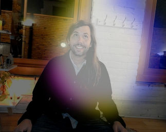 Smiling long-haired person seated on a wooden bench in a cozy urban loft with white painted brick wall, wooden window frames, hanging hooks, potted plant, warm ambient lighting and a purple-yellow lens flare