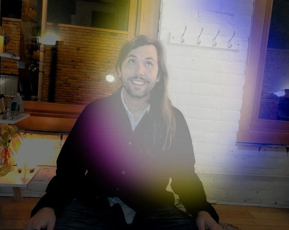 Smiling long-haired person seated on a wooden bench in a cozy urban loft with white painted brick wall, wooden window frames, hanging hooks, potted plant, warm ambient lighting and a purple-yellow lens flare