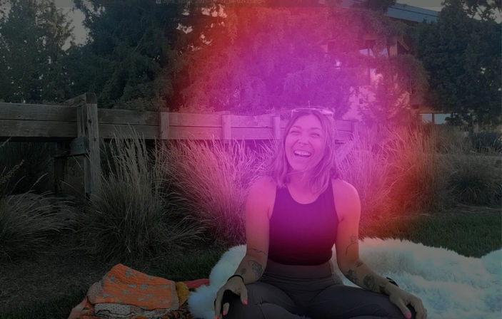 Smiling person with tattoos sitting cross-legged on a blanket in a grassy park by a wooden fence and ornamental grasses, wearing a black tank top and sunglasses on their head, with a vibrant pink-purple light leak effect.