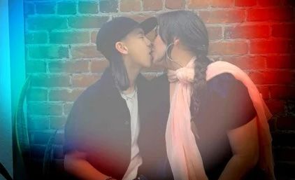 Two people sharing a kiss against an exposed brick wall with a colorful light-leak overlay; one wearing a cap, the other a pink scarf and braid.