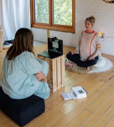 Cozy sunlit home studio with two people doing an online guided meditation — one seated cross-legged on a fluffy rug while a laptop rests on a wooden crate.