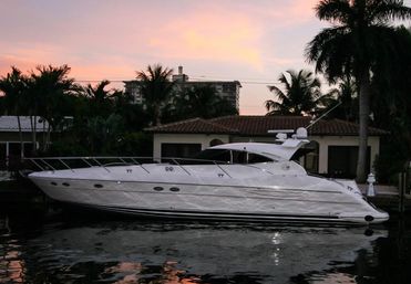 Sleek luxury white motor yacht docked at a tropical waterfront home with palm trees and a colorful sunset reflected in calm canal water.