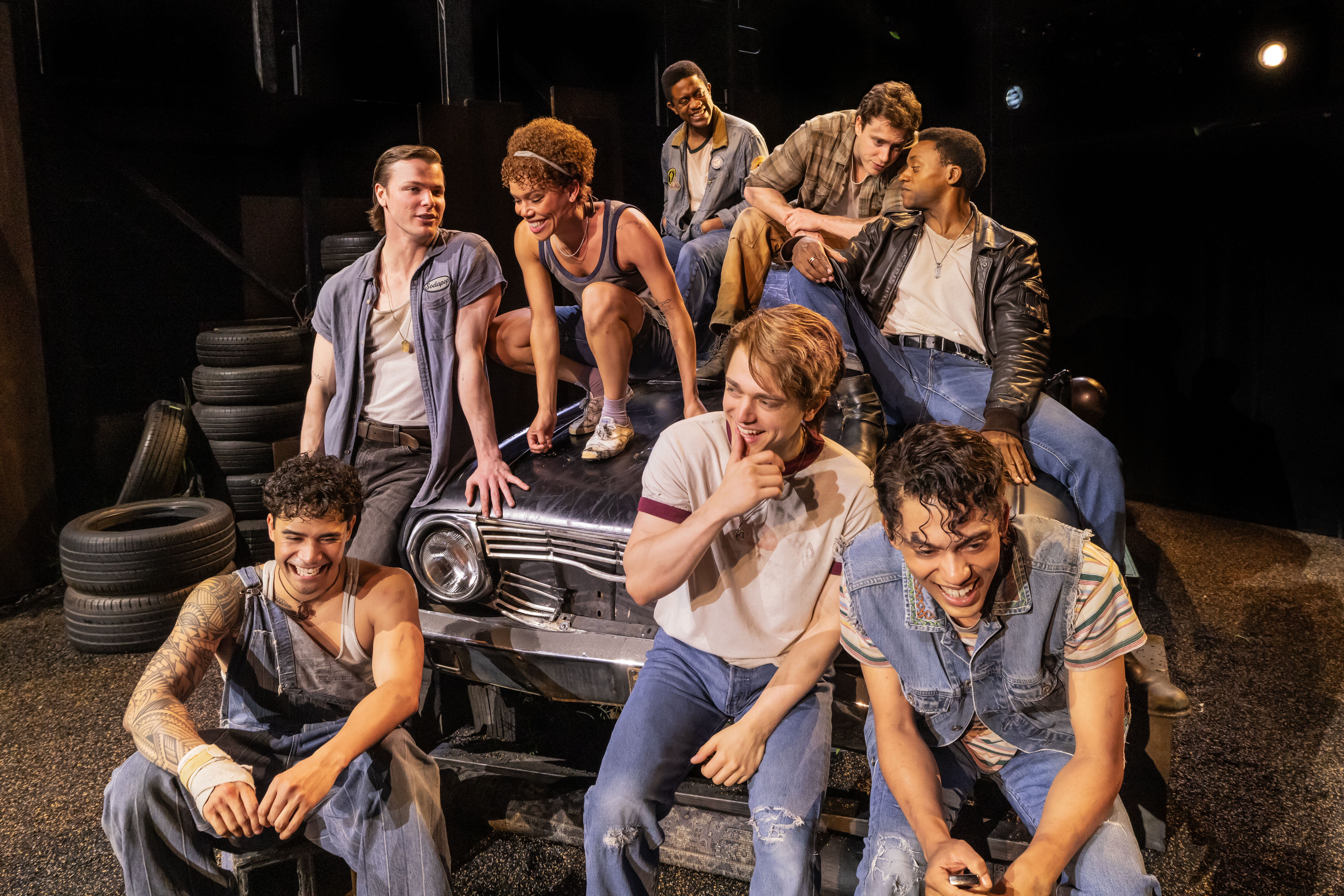 Eight young actors in denim and leather laughing and lounging on a worn car hood amid stacked tires on a dimly lit theater garage set