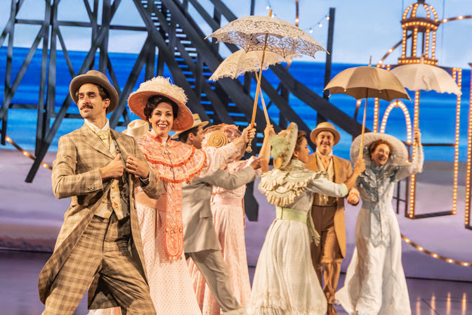 Musical theater ensemble in Edwardian costumes dancing with parasols on a brightly lit seaside pier stage.