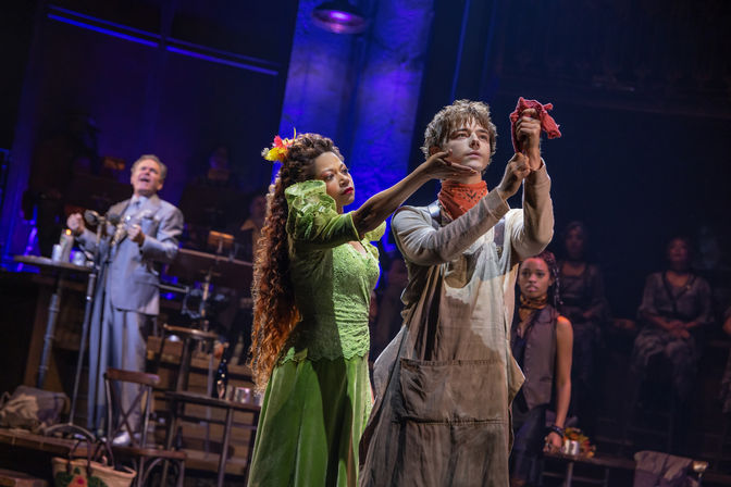 Live theater scene: a woman in a green period dress adjusts a young man in dusty overalls holding a red cloth, framed by purple stage lighting and a background ensemble in a dramatic musical production.