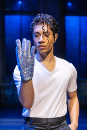 Stage performer in a white tee and black pants inspects a sparkling sequin glove under moody blue stage lighting, dramatic curled wet-look hair.