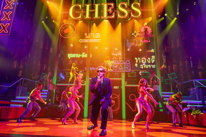 Stage musical scene: lead singer in a suit and sunglasses fronts a neon-lit nightclub set with Thai signage and a large 'CHESS' marquee, dancers and colorful spotlights.