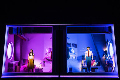 Neon-lit theater stage with two adjacent apartment windows: left room in magenta with a woman standing amid boxes and a fluffy rug; right room in blue with a man in a shirt and tie by a sofa — split-screen modern stage set.