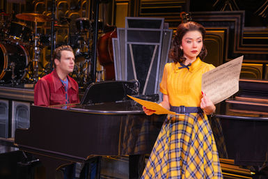 Theater scene: woman in a yellow blouse and yellow-blue plaid skirt holds sheet music by a black grand piano while a man in a red jacket sits at the piano, with drums and a double bass on stage behind them.