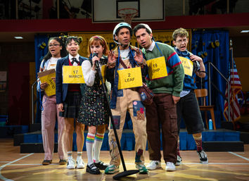 Six teens in colorful outfits and numbered yellow bee-themed placards crowded around a microphone on a school gym stage during a lively spelling-bee performance, with blue curtains and an American flag in the background.