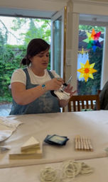 Person in apron demonstrating tie-dye folding and binding with clothespins and rubber bands at a sunlit indoor craft workshop by patio doors with colorful star window decals.