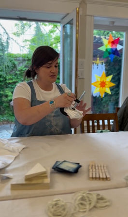 Person in apron demonstrating tie-dye folding and binding with clothespins and rubber bands at a sunlit indoor craft workshop by patio doors with colorful star window decals.