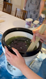 Hands stirring dark green-and-purple fabric dye in a white bucket with wooden paddles on a covered dining table — home DIY tie-dye setup