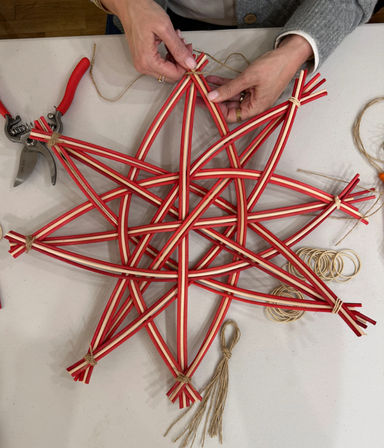 Hands tying twine on a woven red-and-natural straw star wreath, DIY holiday craft on a worktable with rubber bands and red-handled shears
