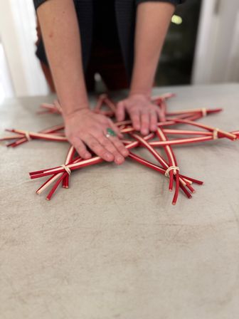 Hands pressing a red-and-natural woven stick star on a table — DIY holiday craft decoration.