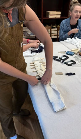 Hands folding a white long-sleeve shirt on a table, securing sections with clothespins and clamps for a DIY tie-dye craft workshop, supplies and other participants visible in the background.