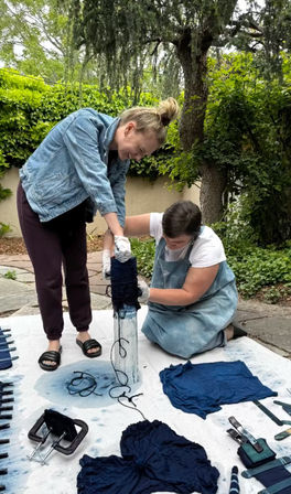 Hands-on indigo tie-dye session on a leafy backyard patio, two people wrapping fabric around a post while indigo-dyed shirts, clamps, and dye-stained tarp surround them.