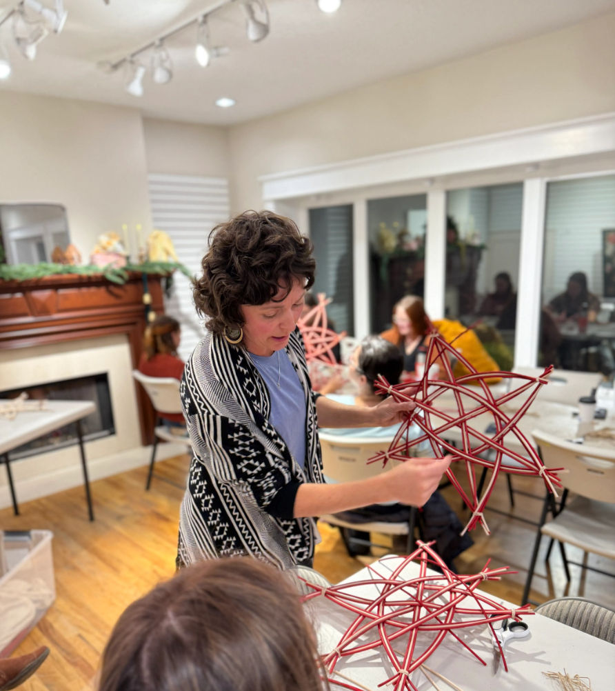 Instructor demonstrating how to weave red twig star ornaments at a cozy indoor community craft workshop with participants seated around tables