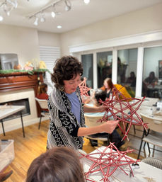 Instructor demonstrating how to weave red twig star ornaments at a cozy indoor community craft workshop with participants seated around tables