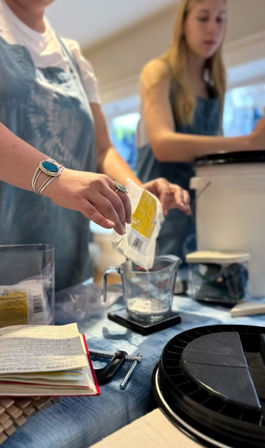 Hands pouring powdered ingredient from a packet into a glass measuring cup on a digital scale, two people in aprons working at a busy kitchen counter with a notebook, clamps and buckets nearby.