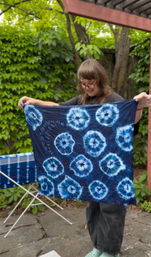 Smiling person holding a square indigo shibori tie-dye cloth with white circular burst patterns, photographed outdoors in a leafy backyard under a wooden pergola
