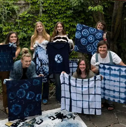 Smiling group of seven on a leafy backyard patio holding indigo shibori tie-dye shirts and fabrics from a DIY dyeing workshop.