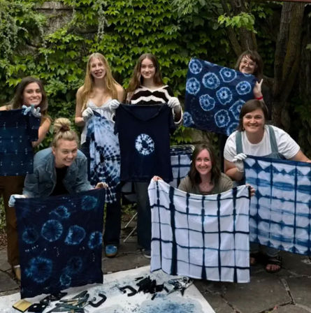 Smiling group of seven on a leafy backyard patio holding indigo shibori tie-dye shirts and fabrics from a DIY dyeing workshop.