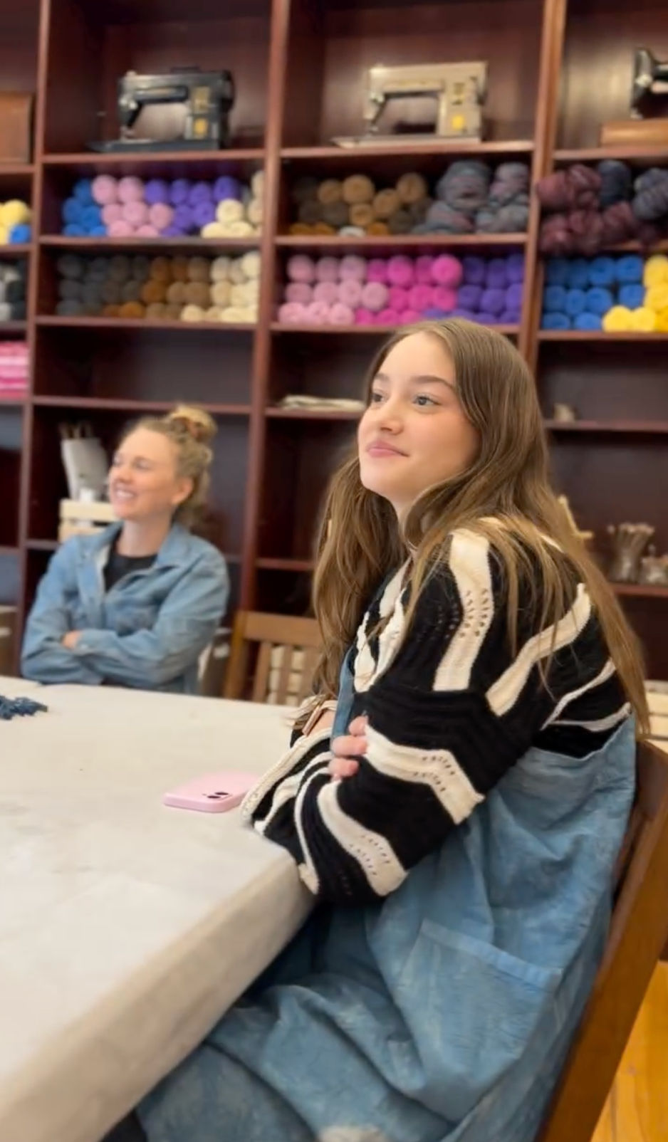 Young woman sitting at a table in a cozy local yarn shop workshop, colorful skeins on wooden shelves and vintage sewing machines in the background.