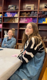 Young woman sitting at a table in a cozy local yarn shop workshop, colorful skeins on wooden shelves and vintage sewing machines in the background.
