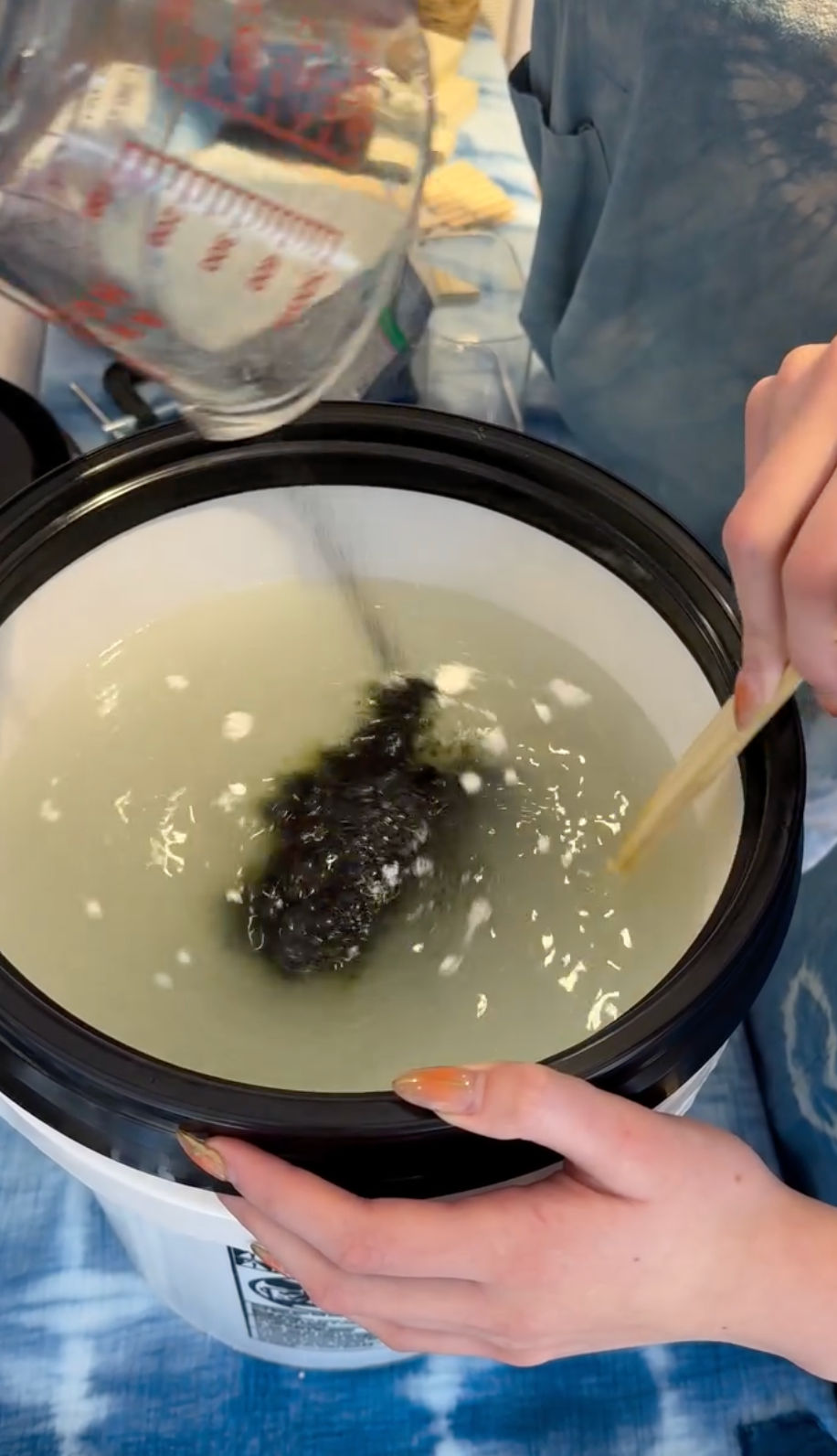 Close-up of hands pouring water from a glass measuring cup into a white bucket while stirring a cloudy dye bath with a wooden stick and a dark fabric bundle, home tie-dye setup.