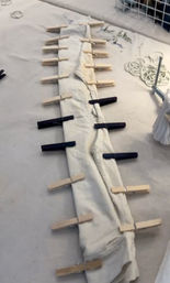 DIY tie-dye prep: long white fabric or pant leg pinned along the edges with alternating wooden and black clothespins on a worktable, with rubber bands and a clamp nearby.