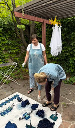 Two people on an ivy-covered backyard patio under a wooden pergola doing indigo tie-dye — clipped blue fabric bundles on a tarp, a folding drying rack and white cloths hanging nearby.