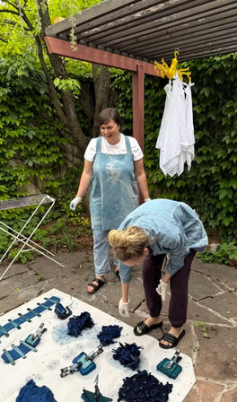 Two people on an ivy-covered backyard patio under a wooden pergola doing indigo tie-dye — clipped blue fabric bundles on a tarp, a folding drying rack and white cloths hanging nearby.
