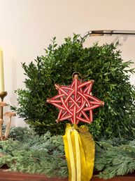 Boxwood holiday wreath on a mantel decorated with a red woven star ornament, yellow ribbon, evergreen garland and candle — festive Christmas home decor.
