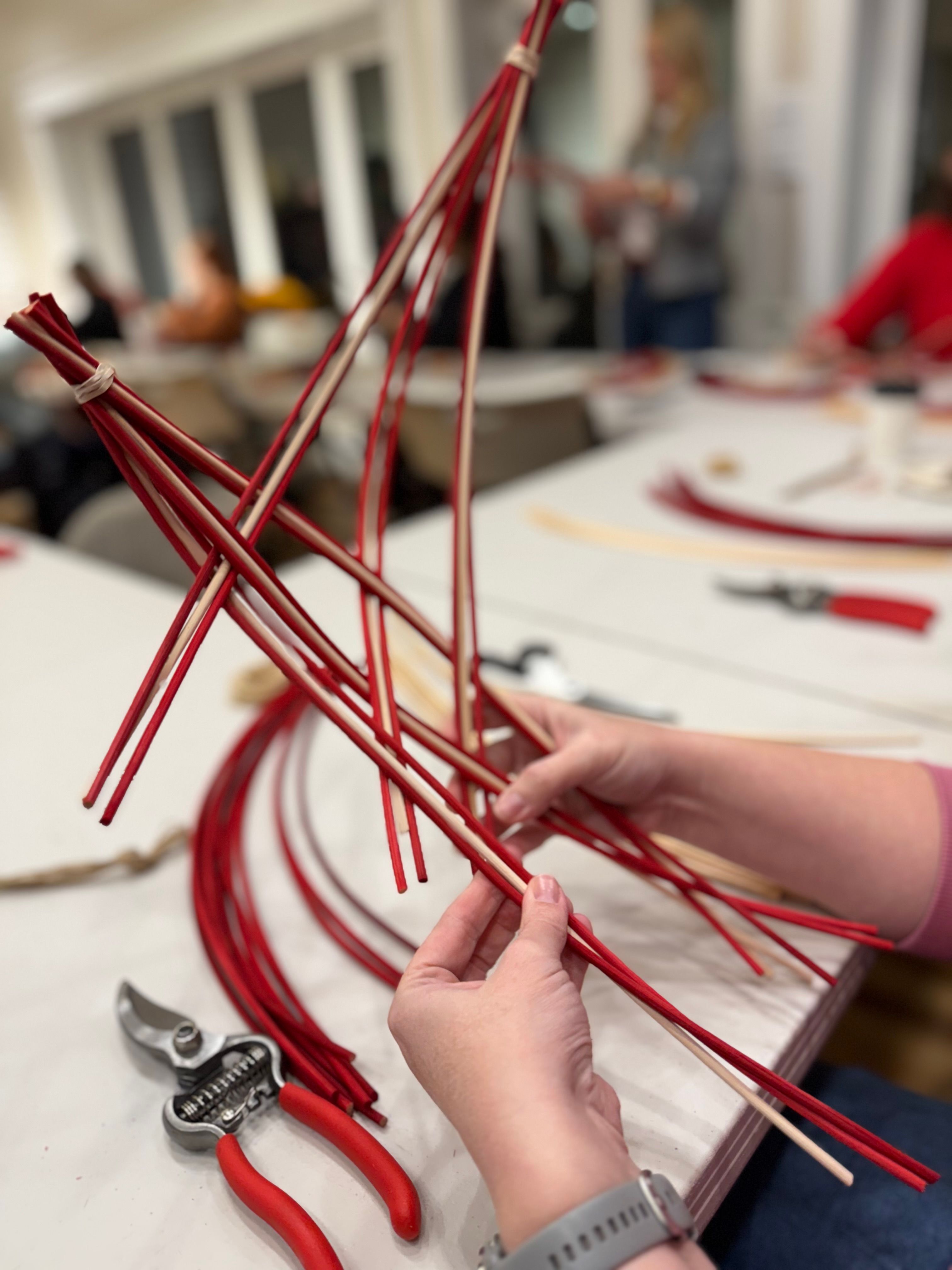 Close-up of hands sculpting bright red willow stems into a decorative frame at an indoor craft workshop table, with red-handled pruning shears and blurred participants in the background.
