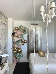 Minimal white bedroom decorated for a celebration with a playful pastel pink and sage green balloon garland, gold metallic accents and a champagne-glass foil balloon by a silver fringe doorway, bed with a gold balloon and white chandelier overhead.